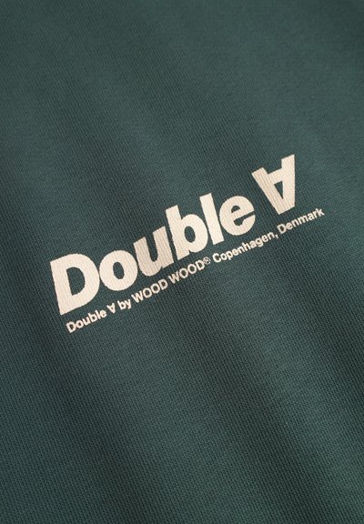 Tissu en coton vert avec du texte blanc : "Double V par WOOD WOOD® Copenhague, Danemark." Texture lisse avec un design respirant.