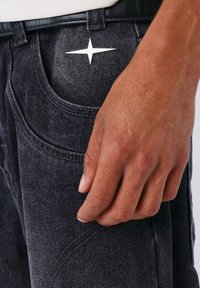Jeans di jeans neri con un design distintivo delle tasche; un accento a forma di stella metallica sul davanti; tessuto testurizzato visibile; mano appoggiata sul materiale.