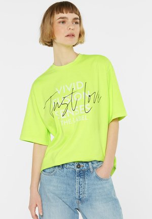 Neon green oversized t-shirt with short sleeves featuring black text, “VIVID FUSION SENSES,” and a casual fit. Paired with light blue jeans.