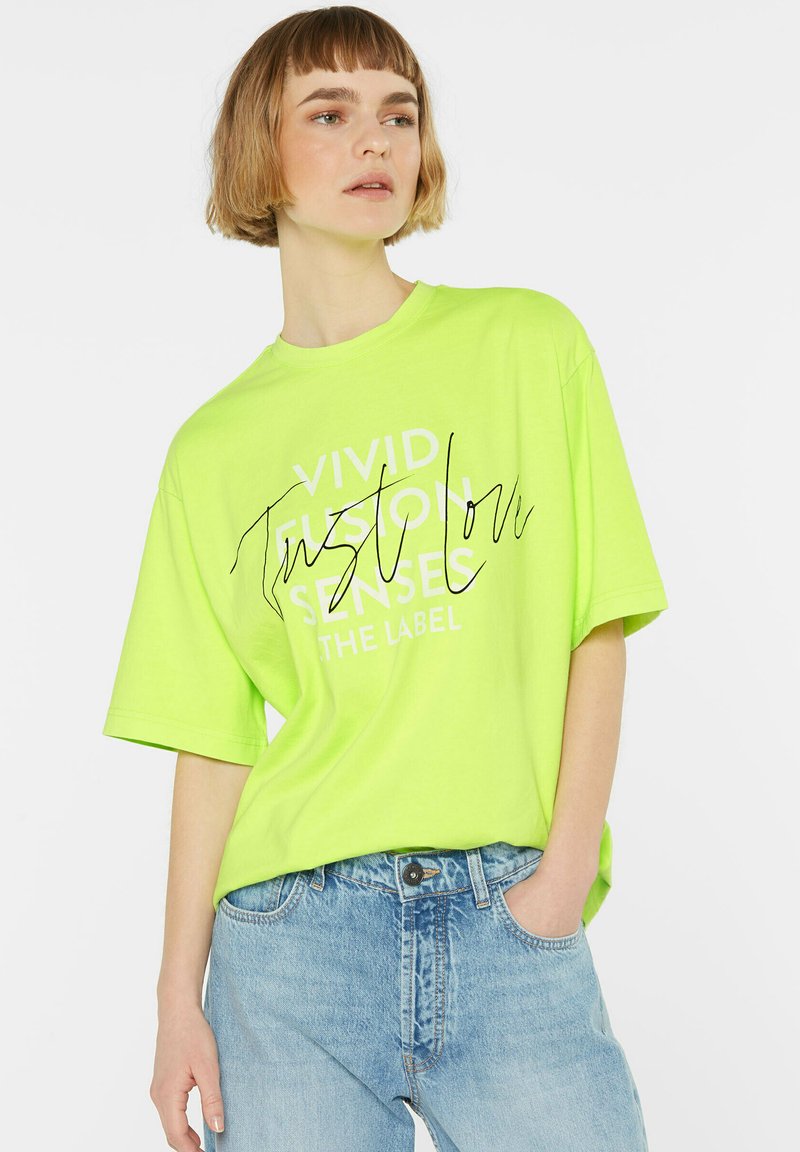 Neongrünes, übergroßes T-Shirt mit kurzen Ärmeln, das schwarzen Text mit der Aufschrift „VIVID FUSION SENSES“ trägt und eine lässige Passform hat. Kombiniert mit hellblauen Jeans.