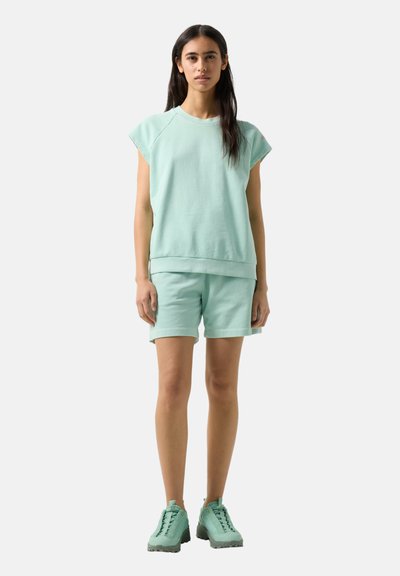 Frau steht, trägt ein passendes ärmelloses mintgrünes Oberteil, Shorts und klobige Turnschuhe, vor einfachem hellem Hintergrund.