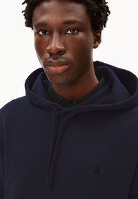 Marineblaue Kapuzenjacke mit Kordelzug, weicher Textur und dezentem Logo-Detail. Hat eine lockere Passform und ist über einem schwarzen Hemd mit Kragen getragen.