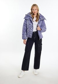 Jachetă pufoasă cu imprimeu de leopard mov, guler oversized, închidere cu fermoar, două buzunare frontale, purtată cu pantaloni cargo negri și teniși albi.