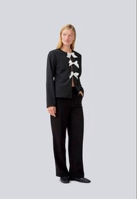 Black jacket with white bow accents down the front, featuring a round neckline and long sleeves, paired with black wide-leg trousers.