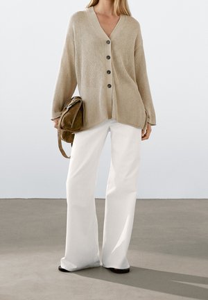 Kobieta w beżowym swetrze na guziki, białych szerokich spodniach, trzymająca brązowy zamszowy plecak na jednolitym tle.