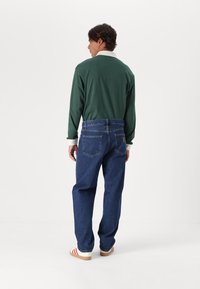 Muž ve tmavě zelené košili s dlouhým rukávem, modrých džínách a bílých teniskách s červenými pruhy, stojící a čelící jednobarevnému bílému pozadí.