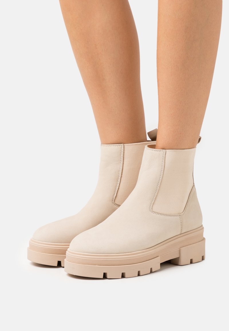 Bottes beige à la cheville avec semelles épaisses et striées portées sur des jambes nues, mettant en valeur un design minimaliste et une texture lisse.