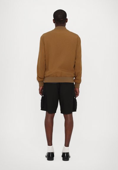 Veste bomber marron avec des poignets et un ourlet côtelés, assortie à un short cargo noir avec des poches latérales, portée avec des chaussettes blanches et des chaussures noires.