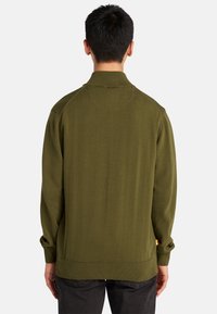 Suéter de punto verde oliva con cuello alto, puños y dobladillo acanalados. Presenta un diseño de espalda sin costuras y una pequeña etiqueta naranja en el lateral.