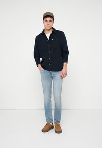 Marinblå skjorta med knappar och två framfickor, i kombination med ljusblå jeans och bruna skor. En mönstrad keps kompletterar outfiten.