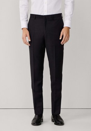 Man wearing tailored black dress pants, white dress shirt, and black dress shoes standing against plain background.