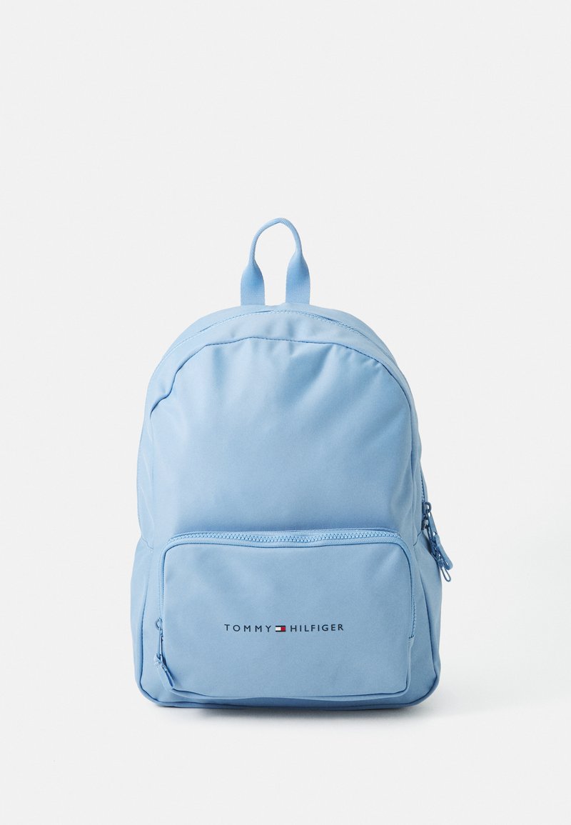 Sac à dos bleu clair en tissu à texture lisse. Dispose d'une poche zippée à l'avant et d'un logo imprimé près du bas. Forme compacte et arrondie.