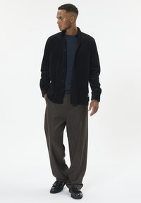 Sort fløjlskjortejakke, blå struktureret sweater under, brune vide bukser og sorte sko. Simpelt design, knaplukning.