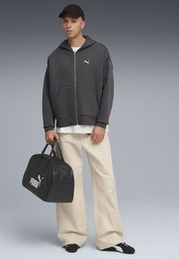 Grijze zip-up hoodie met een kangoeroezak, gecombineerd met beige wijde broek en zwarte sneakers. Draagt een zwarte leren sporttas met een wit logo.