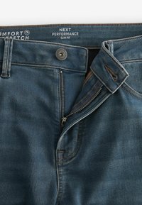 Traperice u tamnoplavoj boji, uskog kroja, s patentnim zatvaračem i kopčanjem na dugme. Čvrsto šavljenje i oznaka s natpisom "Comfort Stretch".
