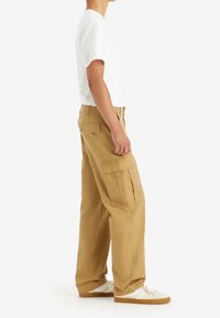 Beżowe spodnie cargo z bocznymi kieszeniami, zwężanymi nogawkami i luźnym krojem, w połączeniu z gładką białą koszulką i białymi trampkami.