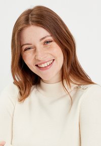 Pull crème avec un col montant côtelé et trois accents de boutons argentés sur l'épaule. Texture douce, silhouette ajustée.