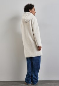 Een capuchon, lange witte jas van een textuurrijk, fluffy materiaal. Draag het over losse, donkerblauwe jeans en sta tegen een lichtgrijze muur.