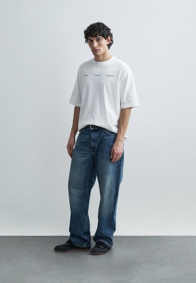 White cotton t-shirt with printed text, paired with blue denim jeans. Minimalist design, relaxed fit, textured fabric. Black shoes with dark soles.