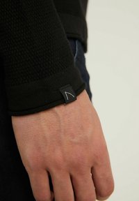 Gros plan d'une main portant une manche texturée noire avec une petite étiquette noire arborant un dessin minimaliste blanc près du poignet.
