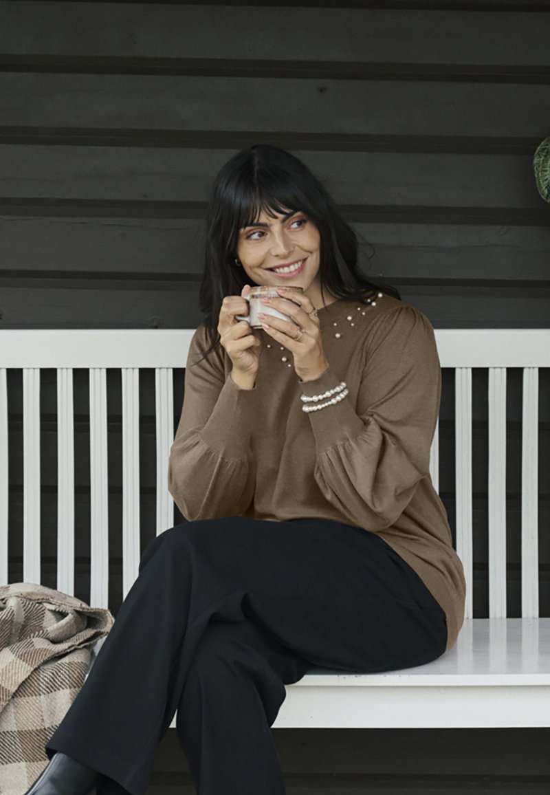 Pull marron texturé avec manches bouffantes et ornements en perles au niveau du col, associé à un pantalon noir, devant un fond en bois.