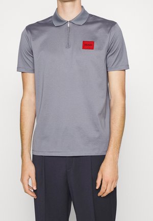 Mann trägt ein hellgraues Poloshirt mit kurzen Ärmeln, vorderem Reißverschluss und rotem "HUGO"-Aufnäher, kombiniert mit dunkler Hose.