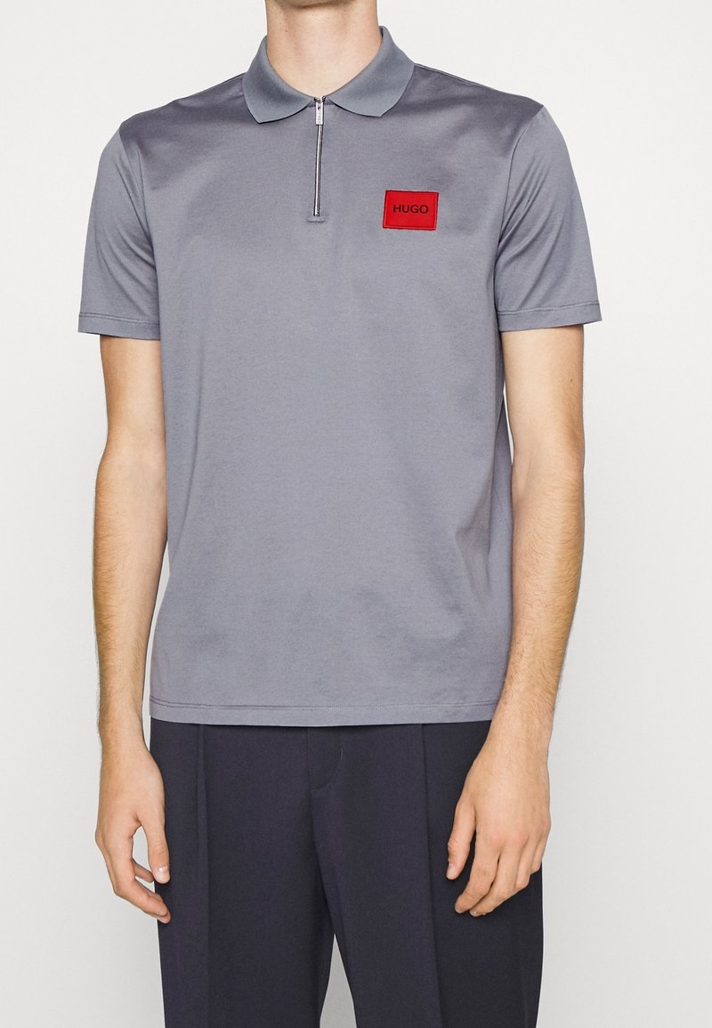Homme portant un polo gris clair à manches courtes avec une fermeture éclair sur le devant et un écusson rouge "HUGO", assorti à un pantalon foncé.