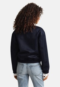 Personne aux cheveux bouclés portant une veste noire et un jean bleu clair, debout dos à un fond blanc.