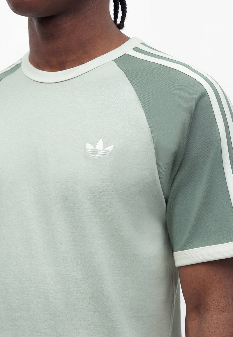 Mand iført lys grøn Adidas T-shirt med hvid kant, tre hvide striber på ærmerne og lille hvid trefoil-logo på brystet.