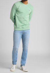 Sudadera verde menta con puños y dobladillo acanalados, que presenta un pequeño logotipo blanco. Combinada con jeans ajustados de color azul claro y zapatillas blancas.