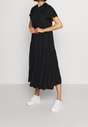 Schwarzes gestuftes Kleid mit Rüschendetails am Ausschnitt, kurzen Ärmeln und Knopfverschluss, kombiniert mit weißen Turnschuhen.