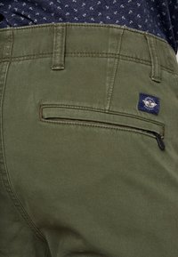 Olijfgroene broek met een gladde structuur, voorzien van een met rits afgesloten achterzak en een klein blauw-wit logo-opdruk nabij de tailleband.