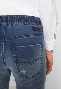 Takana tasa a cilvēka ar zilu džinsu biksēm, kam ir elastīga jostasvieta un neliela plīsuma aizmugures kabatā, apģērbs ir balts garām piedurknēm. 