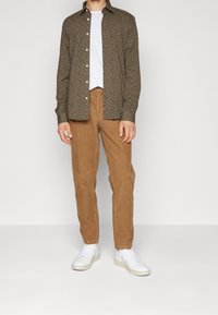 Camicia a maniche lunghe a righe marroni, t-shirt bianca a girocollo, pantaloni in velluto a coste beige e sneakers bianche. La camicia ha una vestibilità slim con colletto.