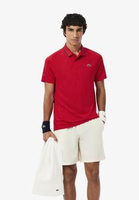 Punainen lyhythihainen polo, jossa on jäsennelty kuvio, kontrastivärinen laivastonsininen kaulus, Lacosten logo, yhdistetty valkoisiin shortseihin ja pyyhkeeseen.