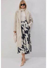 Beige wollen jas met grote revers, crème coltrui, zwart-crème patroon rok, beige clutch en nude puntige hakken.