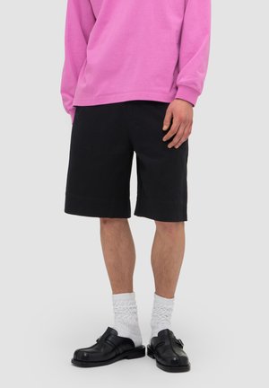 Persoon draagt zwarte knielange shorts, witte gestructureerde sokken, zwarte leren gespschoenen en een roze longsleeve tegen een grijze achtergrond.