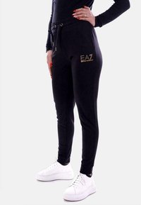 Zwarte joggingbroek van zacht materiaal, met zijzakken, een elastische tailleband met trekkoord en een gouden logo op de heup.