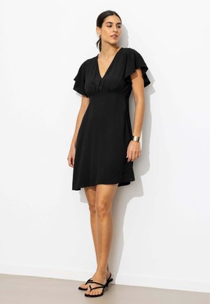 Femme debout contre un mur blanc, portant une robe noire courte à manches volantes et des sandales à talons ouvertes noires, bracelet en or au poignet.