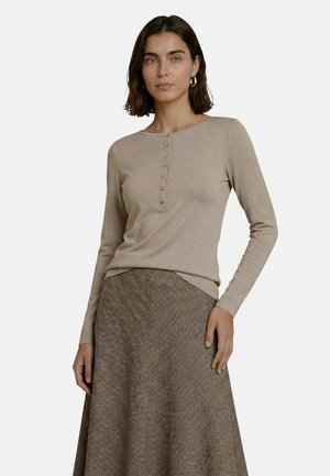 Femme portant un haut beige à manches longues avec des boutons et une jupe marron texturée coupe trapèze, debout devant un fond clair uni.