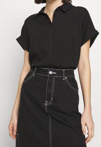 Chemise noire à manches courtes avec col et boutons, associée à une jupe en jean noire taille haute avec des surpiqûres blanches contrastantes.