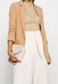 Beige bleiseri, ribbineuloksinen vaaleanruskea poolopaita, valkoiset korkeavyötäröiset housut, joissa on solmittava vyö, ja vaaleanbeige clutch-laukku läpällisellä suljinnalla.