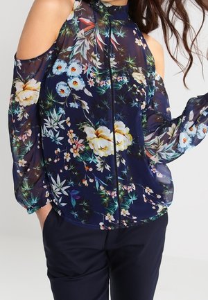 Marineblå blomstret kold skulder-bluse med gennemsigtige lange ærmer kombineret med mørke bukser på en person.