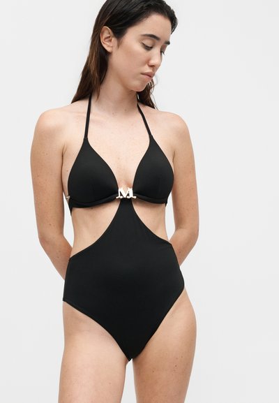 Γυναίκα φοράει μαύρο ολόσωμο μαγιό με halter λαιμόκοψη και ασημένιο διακοσμητικό "M" στο κέντρο, κοιτάζοντας προς τα κάτω με τα χέρια πίσω από την πλάτη.