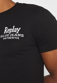 Czarna koszulka z krótkim rękawem wykonana z miękkiego bawełnianego materiału. Z przodu znajduje się biały logo z napisem "Replay Blue Jeans Authentic."