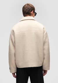 Kurtka polarowa w jasnym beżu o luźnym kroju, z dużym kołnierzem i strukturalnym wykończeniem. Posiada opadające ramiona i prosty dół.