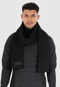 Black knit scarf with ribbed texture, featuring a solid color and a small text detail at one end, worn over a black jacket.