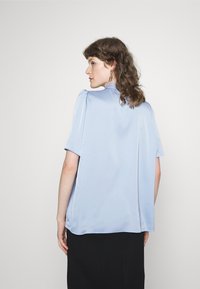 Blusa em cetim azul claro com mangas curtas e um corte solto. Apresenta preguinhas discretas nos ombros e uma textura lisa, ligeiramente reflexiva.