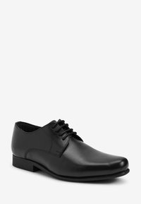 Chaussure habillée en cuir noir avec texture lisse, bout arrondi, design à lacets et détails de couture minimalistes. Dotée d'un talon bas et plat.