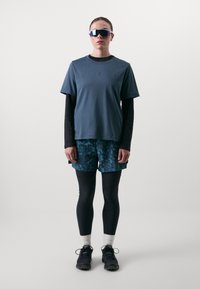 Sportlik riietus, mis sisaldab tumedat sinist T-särki, mustrilisi lühikesi pükse sinistes toonides, musti retuuse ja peegelprille.